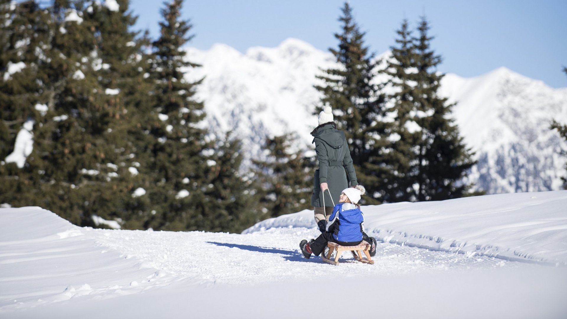 Das Skigebiet Ladurns und andere Winter-Highlights Frau zieht Kind auf Schlitten durch verschneite Winterlandschaft mit Tannen