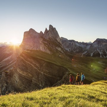 Gudruns Blog: alles über Sterzing, übers Wandern, über Tirol Drei Wanderer auf grünem Hügel mit Sonnenuntergang und Bergpanorama