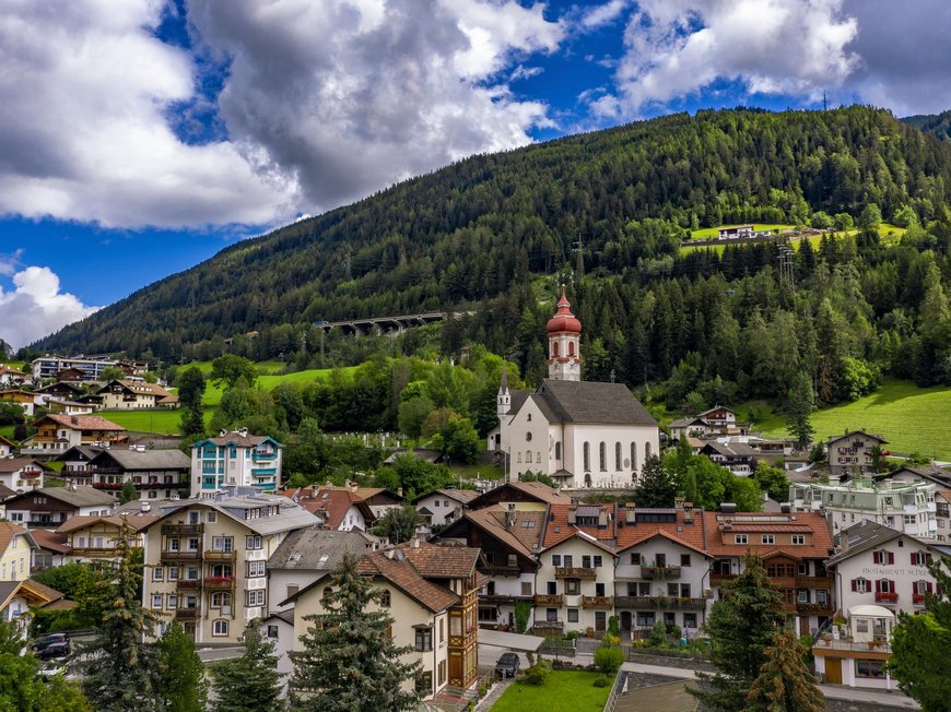 Hotel nahe Pflerschtal: Lage und Anreise Blick auf ein Dorf mit Kirche vor bewaldetem Hügel unter bewölktem Himmel