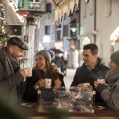 Das Skigebiet Ladurns und andere Winter-Highlights Vier Menschen trinken Heißgetränke an einem beheizten Stehtisch draußen im Winter
