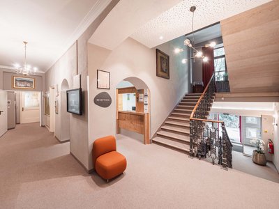 Hotel in Gossensass: welcome to Gudrun! Helles Hotelfoyer mit Rezeption, Treppe und orangefarbenem Sessel.
